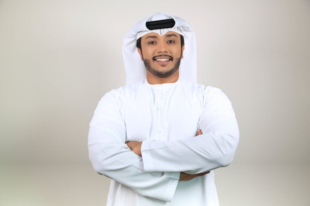 Confident Middle Eastern man smiling in traditional attire with crossed arms, studio portrait.