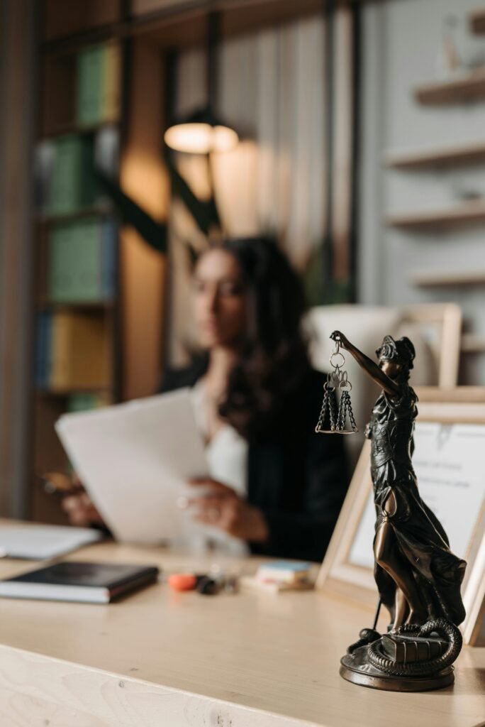 Close-up of a justice figurine on desk with blurred female lawyer in office setting.